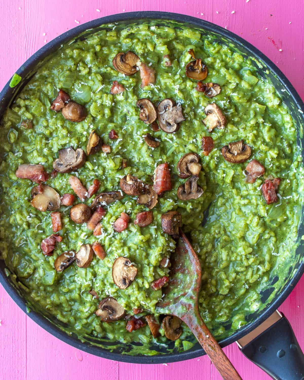 A large pan with green risotto topped with bacon pieces, mushrooms and parmesan cheese.
