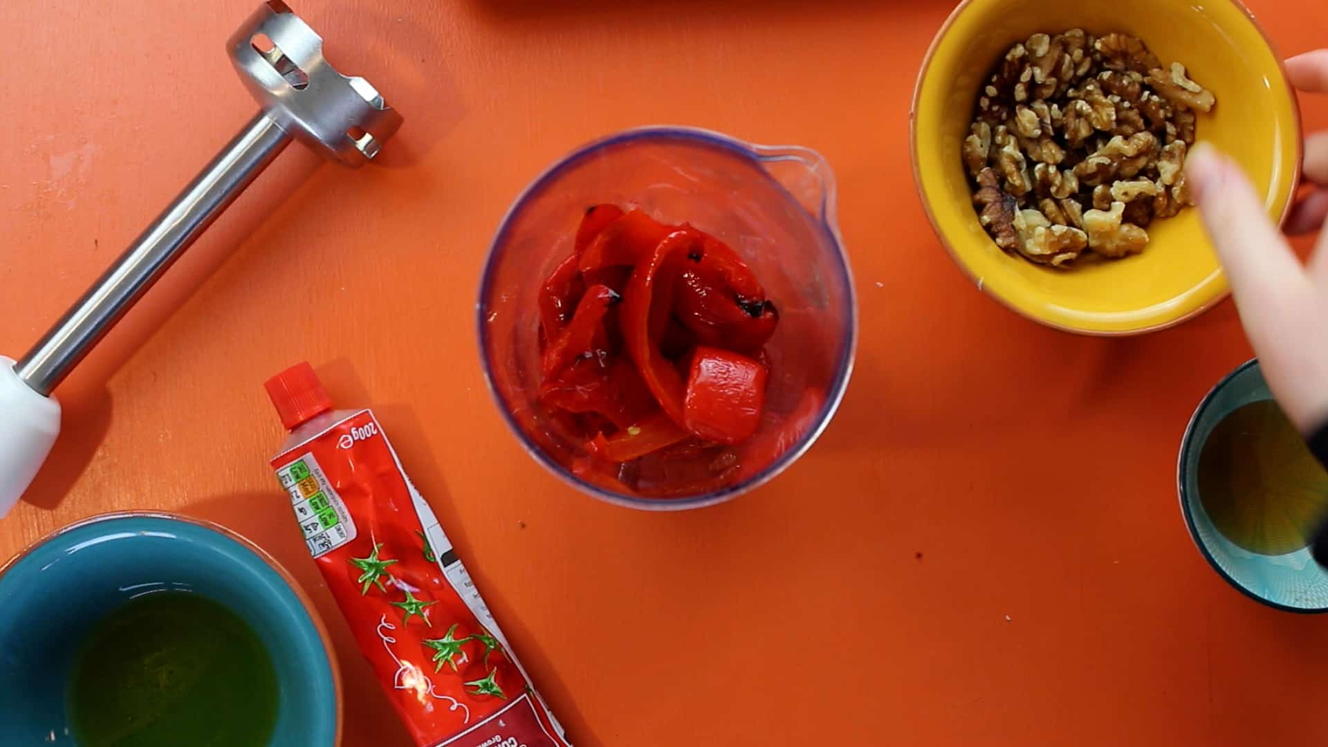 Roasted red peppers in a blended cup with walnuts in a bowl and tomato paste in tube and hand blender on an orange background.