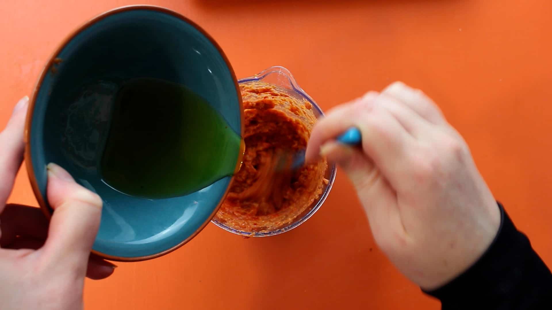 Olive oil being added into the blended sauce Roasted red peppers in a blended cup with walnuts in a bowl and tomato paste in tube and hand blender on an orange background.