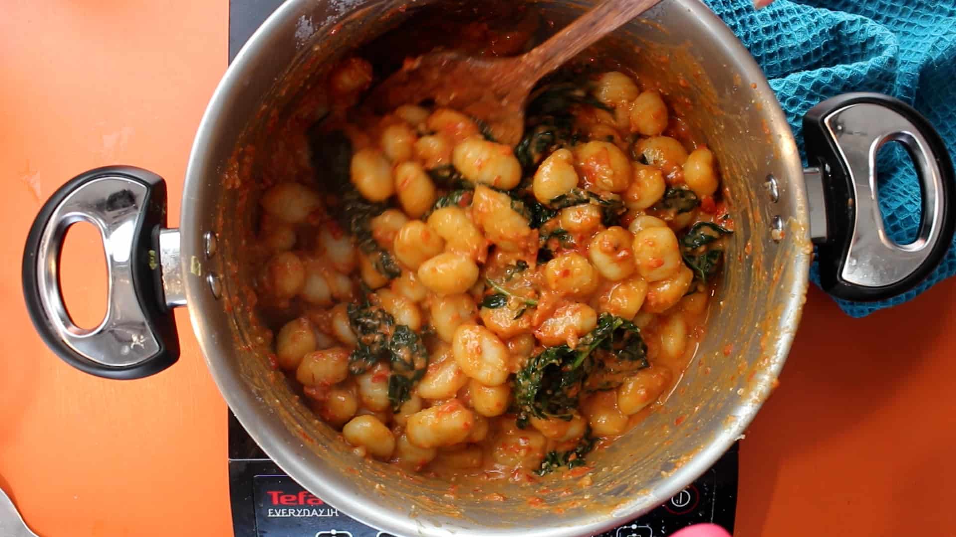 Sauce added to the cooked gnocchi and cavolo nero in a large saucepan and mixed with a wooden spoon on stove on an orange background.