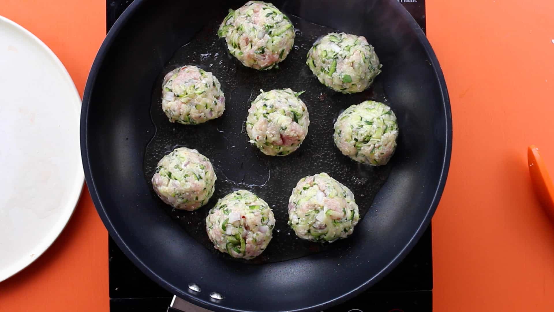 8 meatballs in large frying pan on stove on an orange background with a white plate at side.