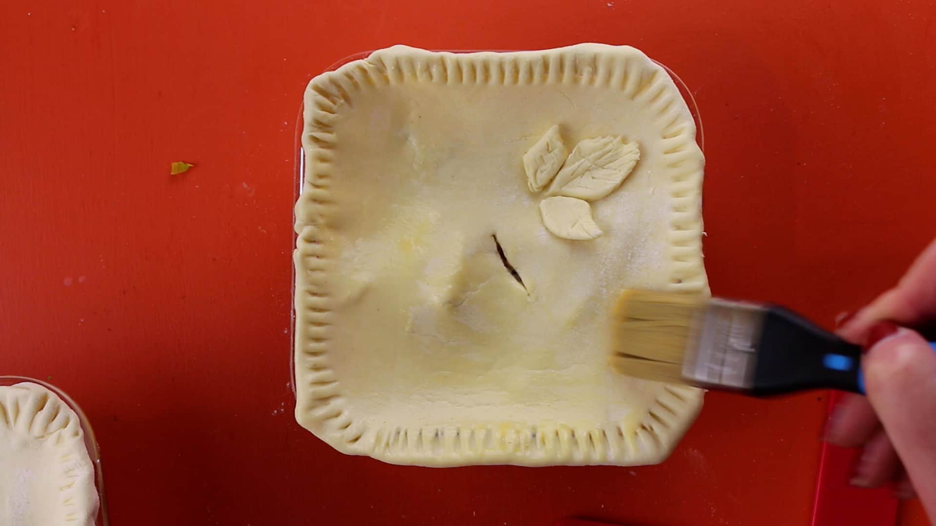 Pastry top added to a square meal prep container, decorated with 3 leaves and brushed with egg wash on an orange background.