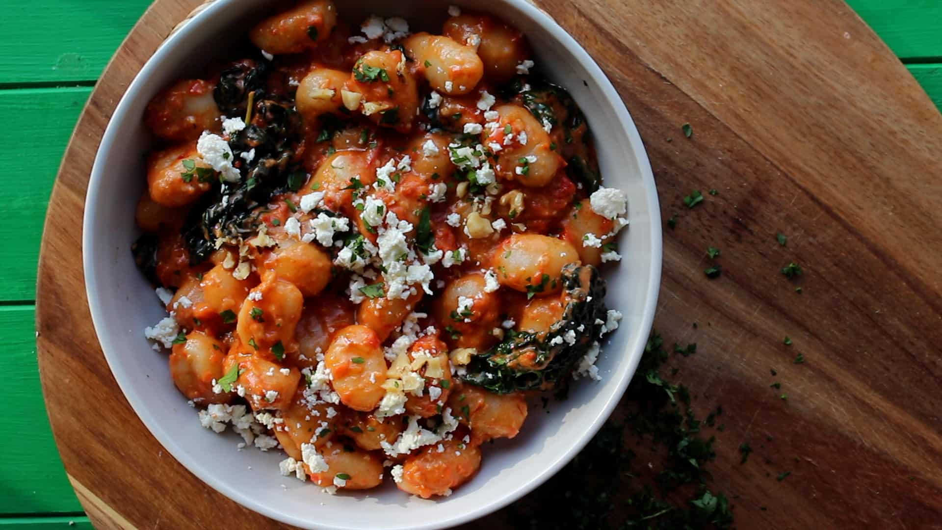 Gnocchi served in a white dish with cavolo nero topped with crumbled feta and walnuts on a wooden board.