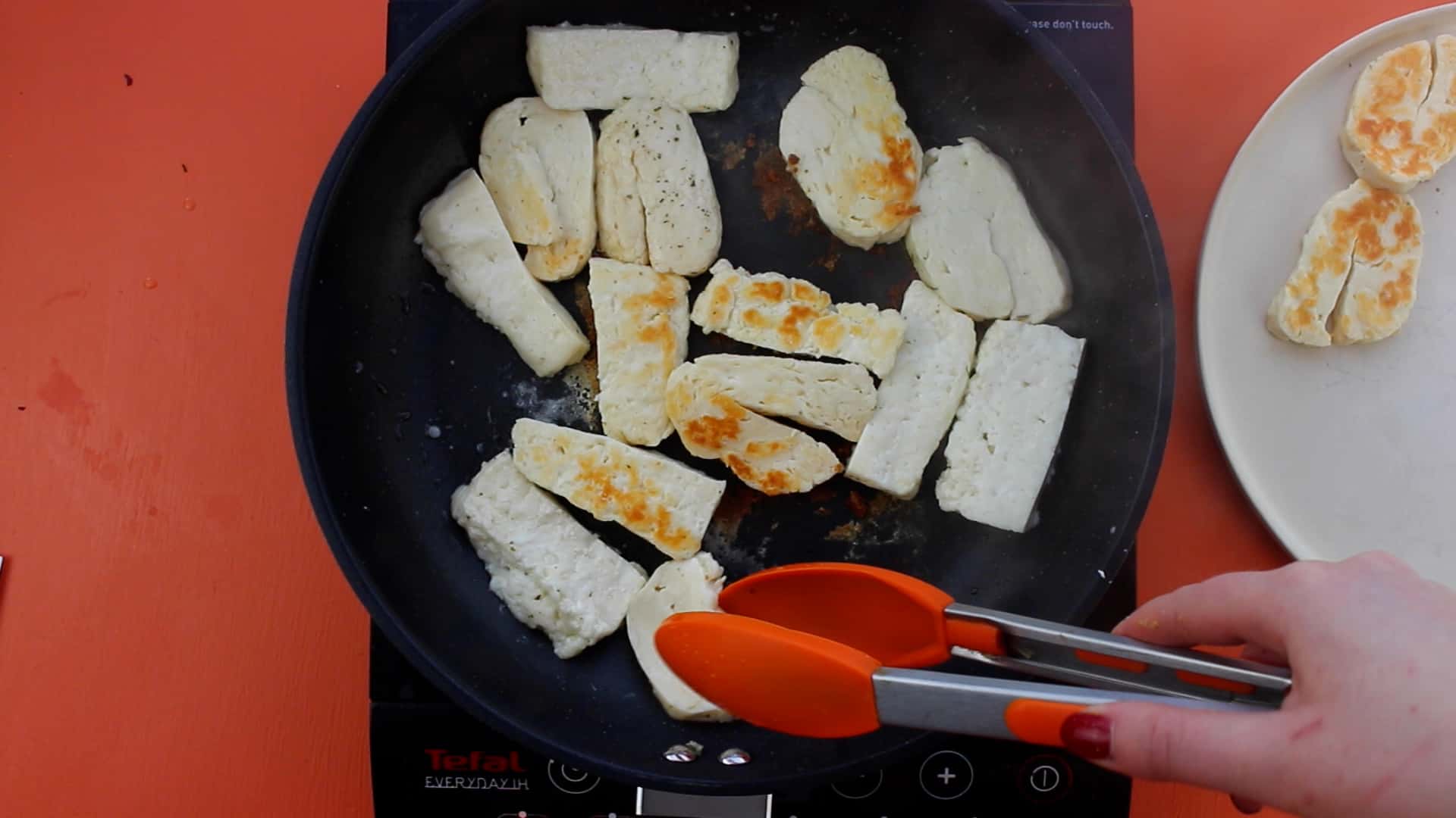 Halloumi being fried in pan until golden brown and then removed from pan with tongs onto a plate.