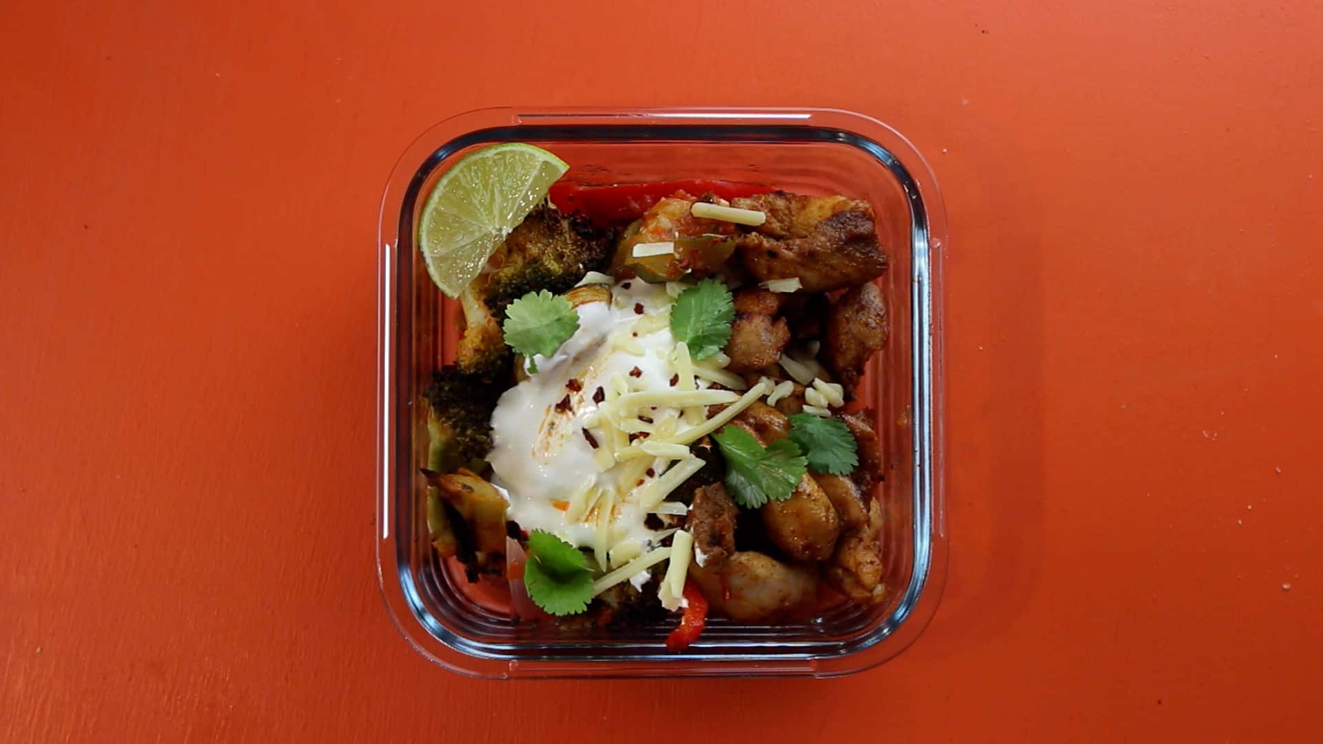 Chicken with sour cream, grated cheese, fresh coriander and wedge of lime in a meal prep container on an orange background.
