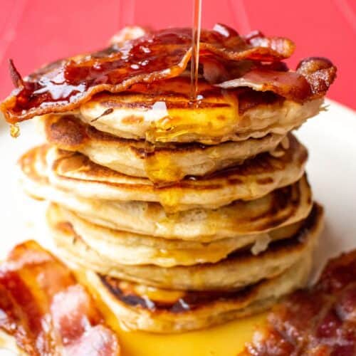 Loads of pancakes piled on top of each other and topped with honey and bacon on a plate with a pink background.