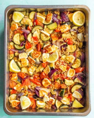 Roasted Mediterranean Vegetables With Halloumi and Baked Orzo with red onions, tomatoes and courgettes with lemon wedges on a baking tray.
