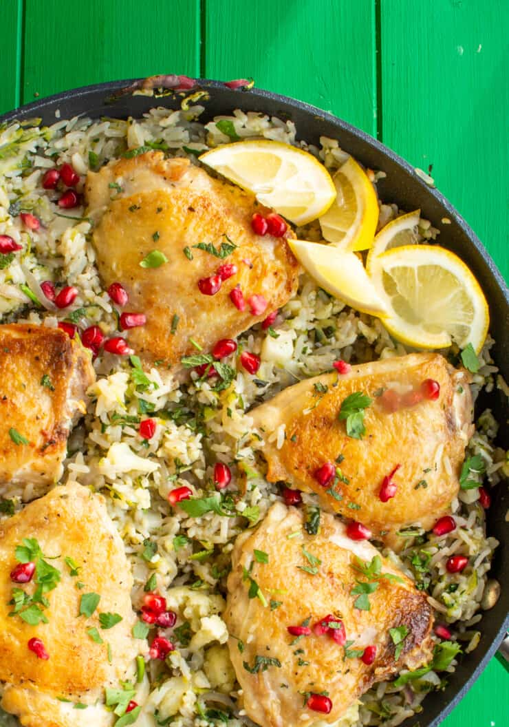 Lebanese Inspired Chicken Pilaf in a pan with rice, 5 browned chicken thighs with mint, parsley and pomegranate seeds on a green background.