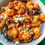Close up of Walnut Romesco Gnocchi, cavolo nero, a tomatoey red pepper sauce topped with crumbled feta, walnuts and parsley in a white bowl.