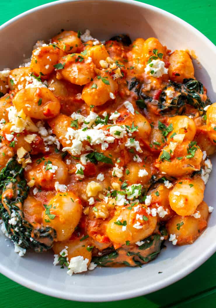 Close up of Walnut Romesco Gnocchi, cavolo nero, a tomatoey red pepper sauce topped with crumbled feta, walnuts and parsley in a white bowl.