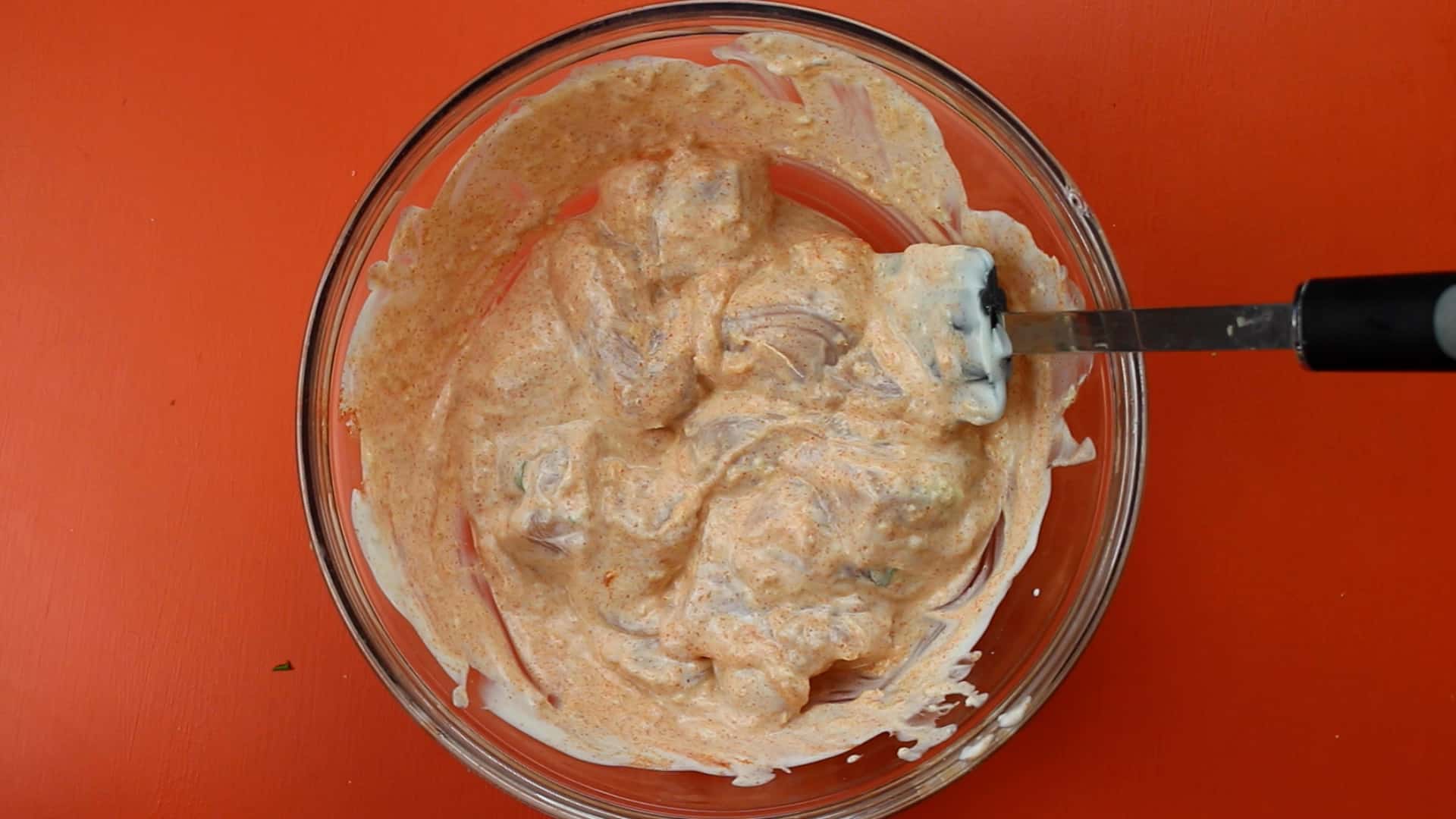 Chicken pieces in a glass bowl with yogurt and spices mixed together with a spatula on an orange background.