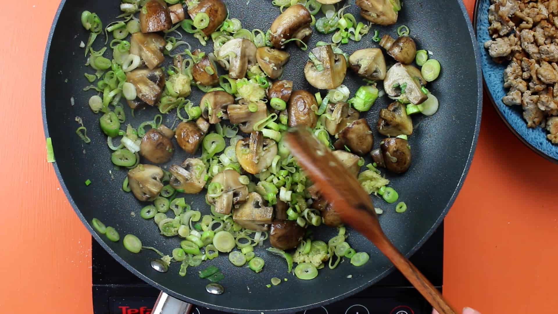Ginger and spring onions added to the mushrooms in pan mixed with a wooden spoon.