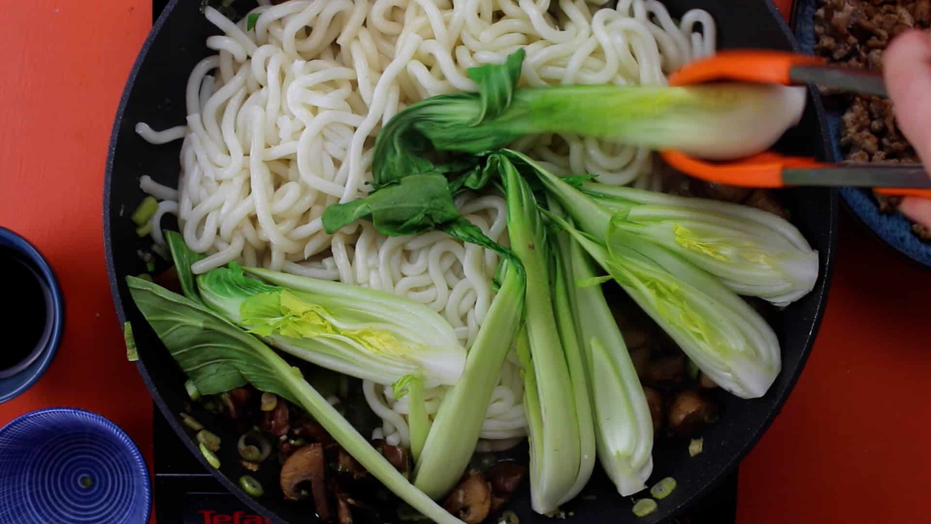 Pak choi slices over the udon noodles in the pan with mushrooms and spring onions with orange tongs moving the pak choi.