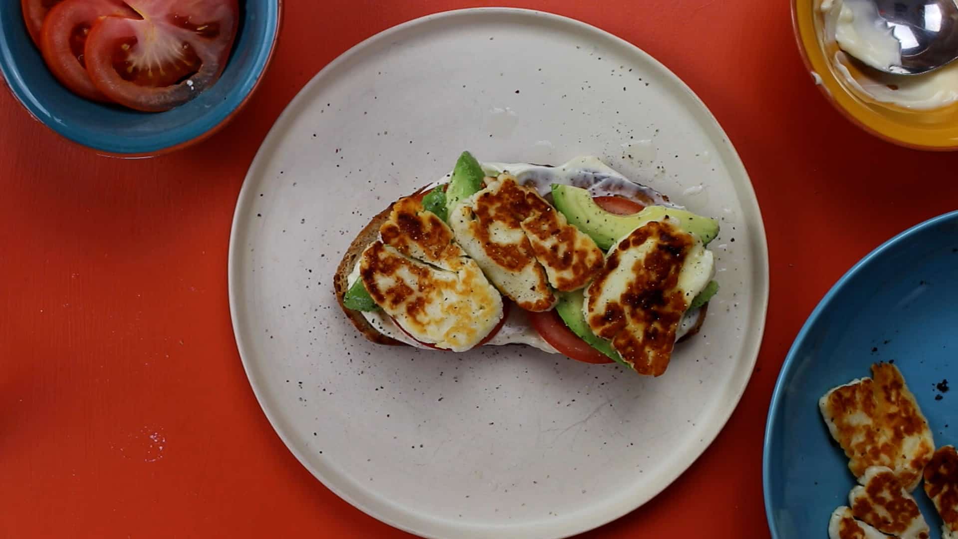 Avocado, tomato, mayonnaise and halloumi on a piece of sourdough on a plate with tomatoes, halloumi and mayonnaise in separate bowls.
