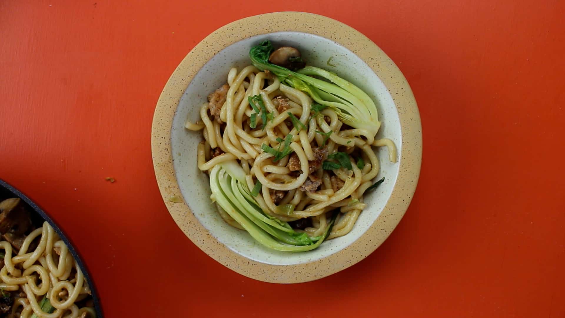 Stir-fry Pork Udon Noodles  with pak choi  and spring onion slices served in a dish on an orange background.