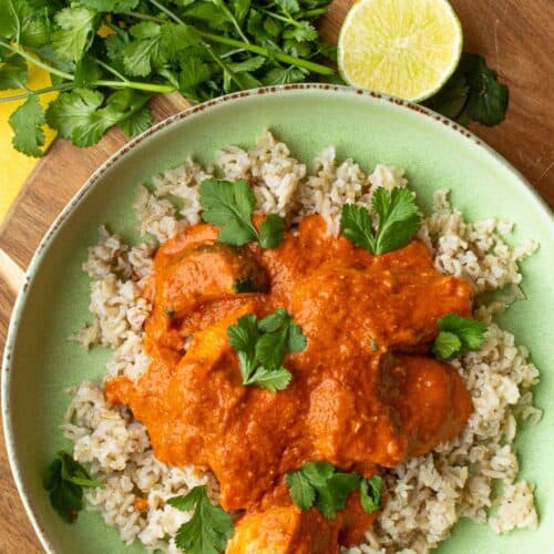 Chicken tikka masala with brown rice and coriander in a green bowl on a wooden board with sliced lime and fresh coriander.