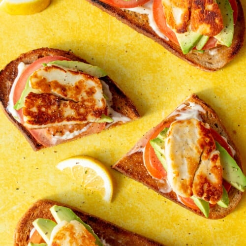 Halloumi and Avocado Sourdough Toast slices with avocado, tomato, mayonnaise topped with golden browned halloumi on a yellow background.