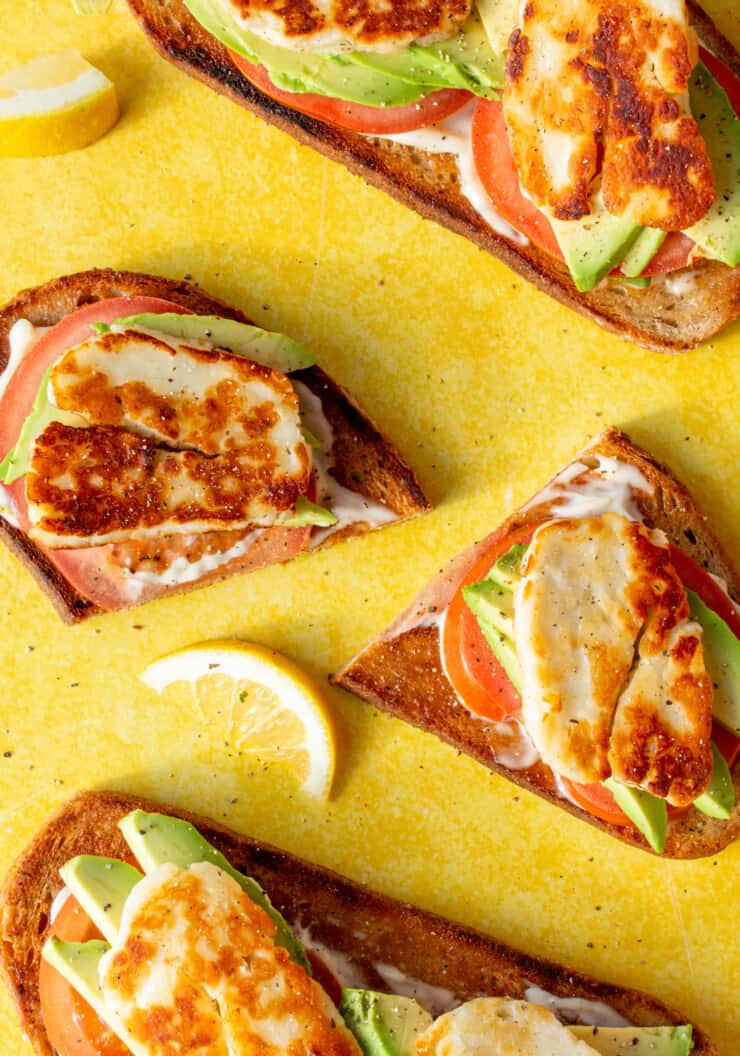 Halloumi and Avocado Sourdough Toast slices with avocado, tomato, mayonnaise topped with golden browned halloumi on a yellow background.