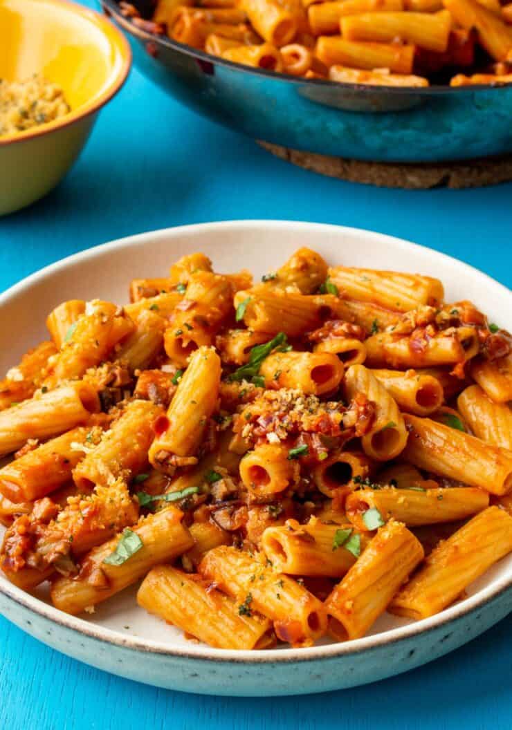 A bowl of Mushroom Rigatoni pasta with breadcrumb topping and fresh basil with a small bowl of breadcrumbs and the pan in partial view.