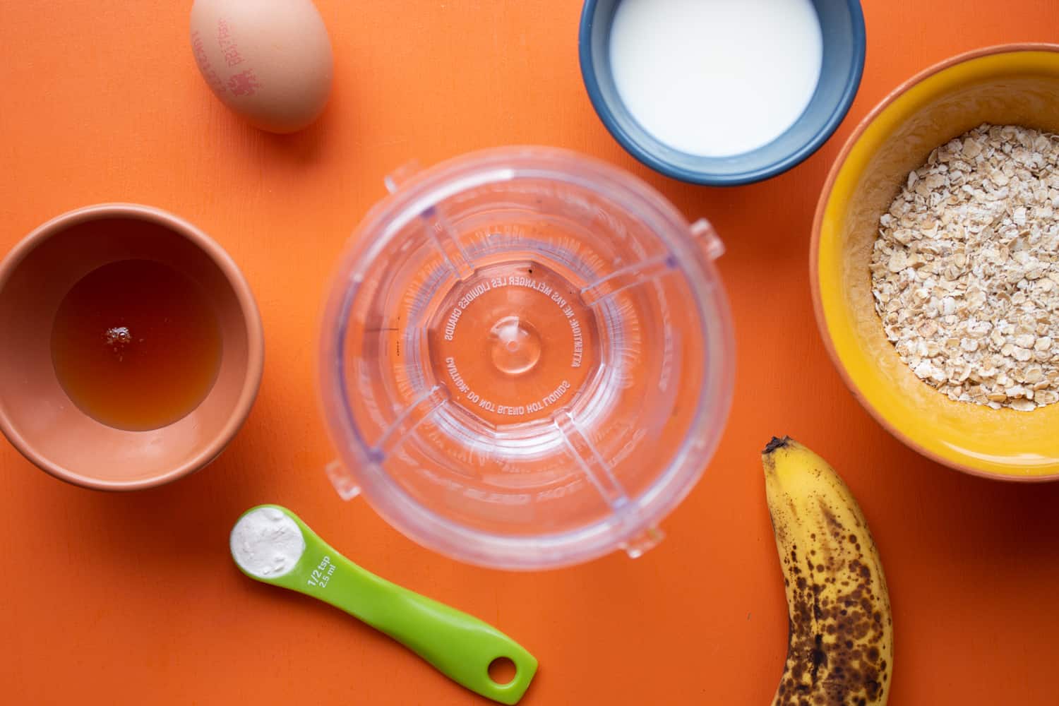 Ingredients including honey, banana, eggs, oats and milk around a blender cup on an orange background.
