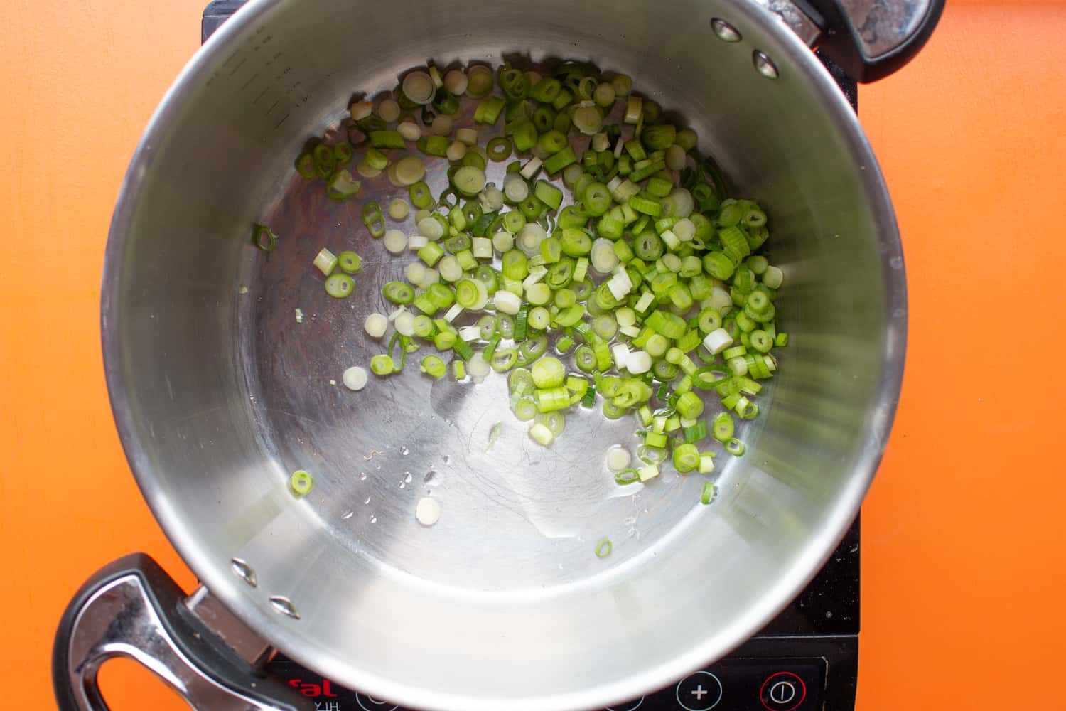 Chopped spring onion in large saucepan with 2 handles on cooker on an orange background.