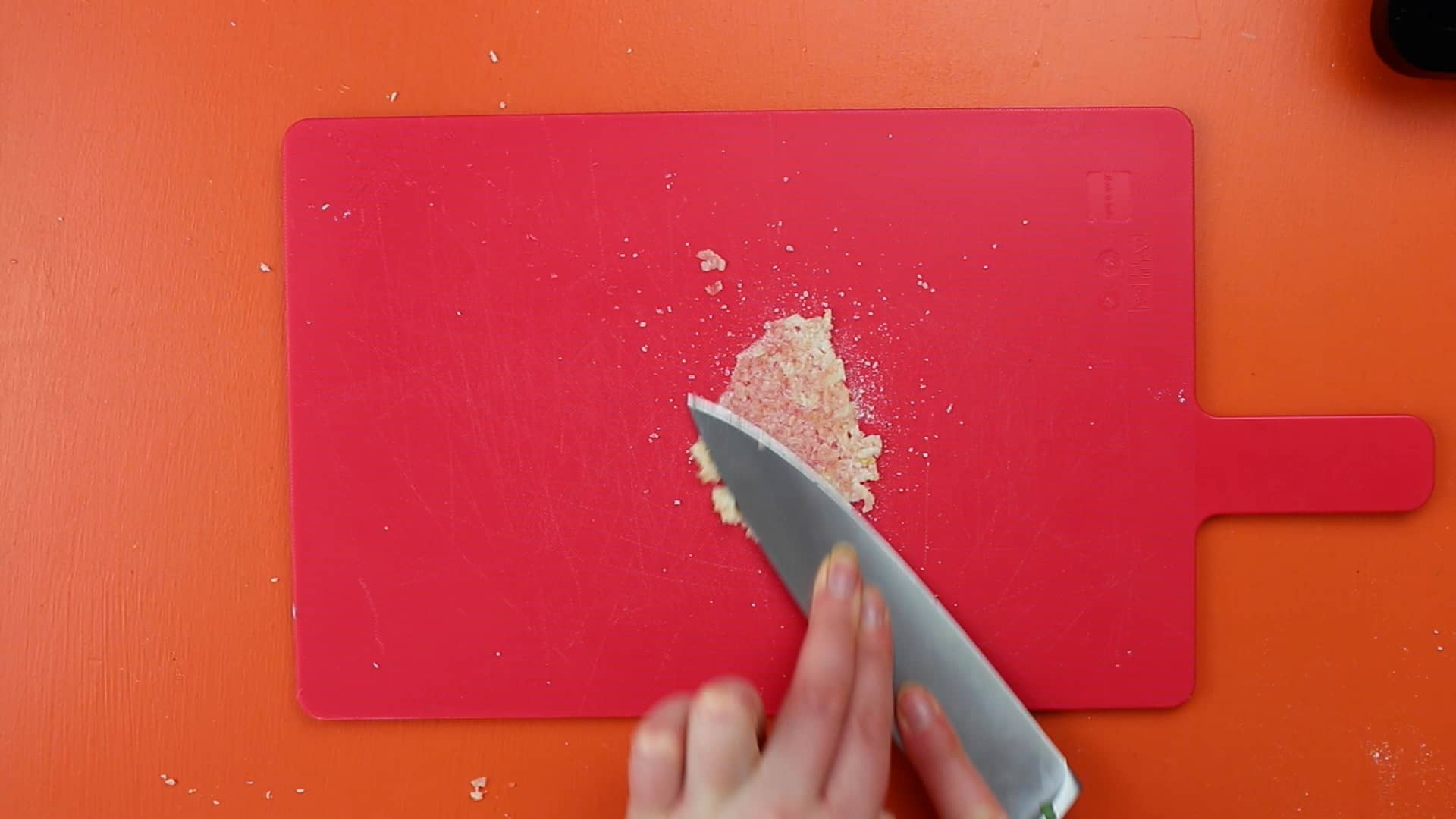 Garlic being pressed with knife on a pink chopping board on an orange background.