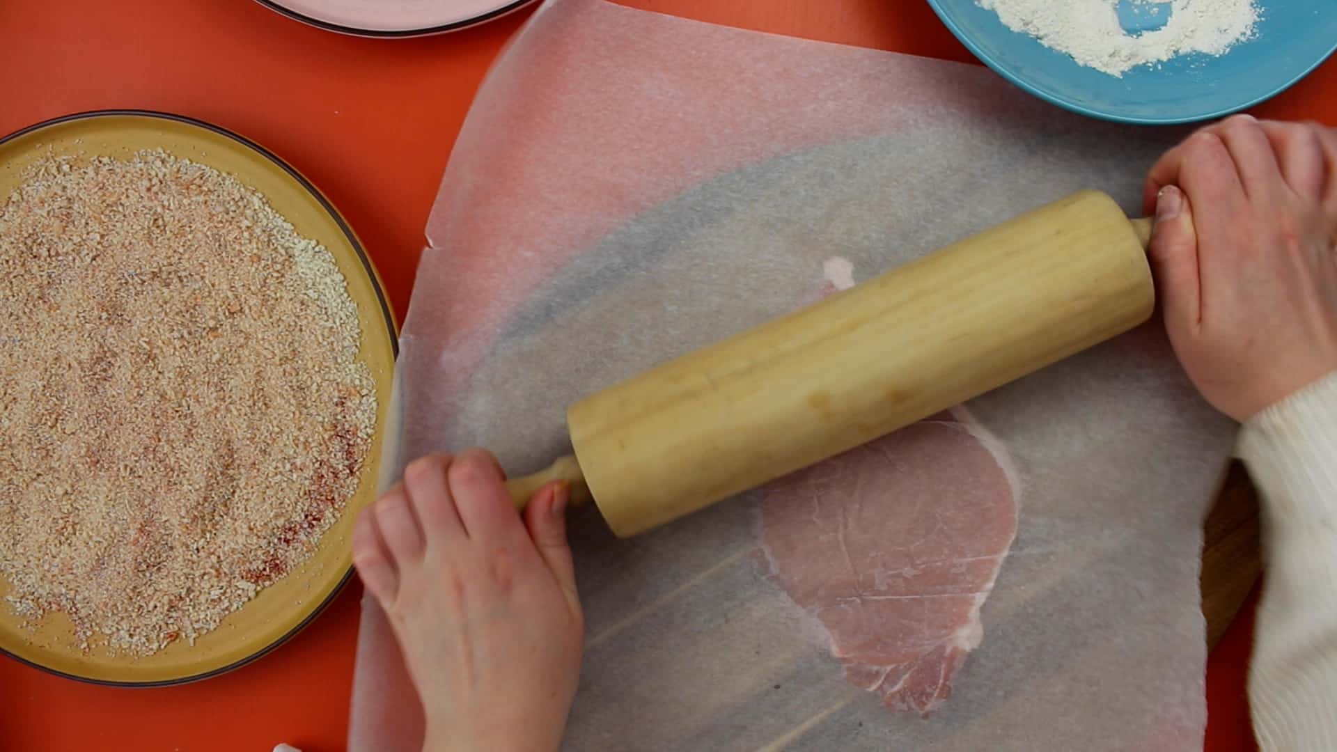 Pork with grease proof paper over the top being pounded with rolling pin with breadcrumbs and flour on separate plates.