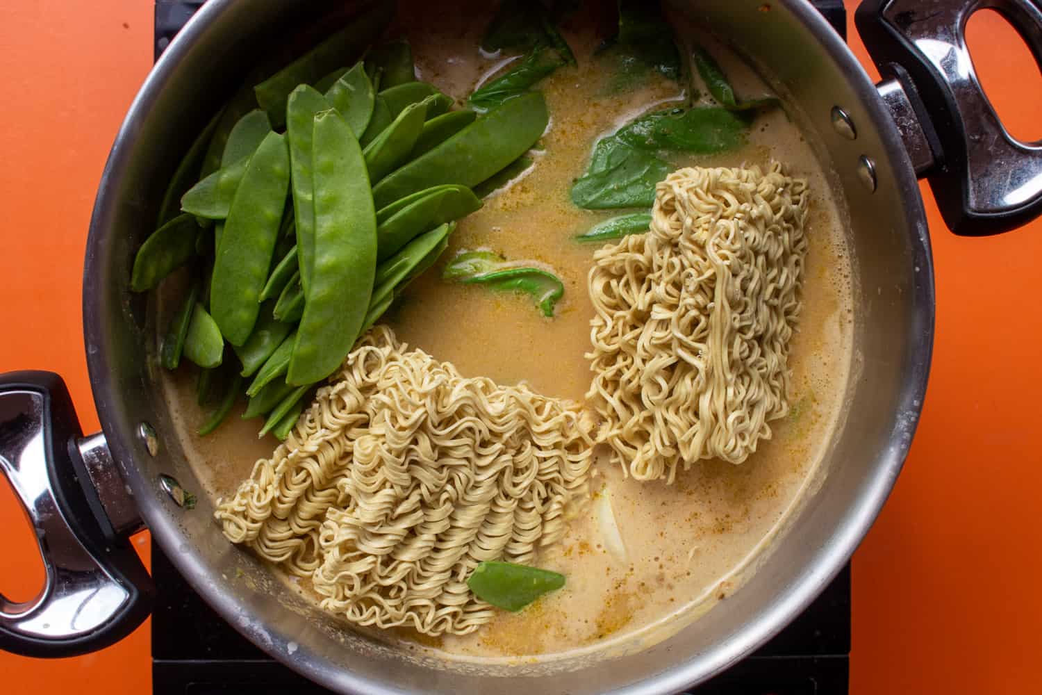 Dry noodles and mange tout added to the ramen in a large saucepan with 2 handles on a stove on an orange background.