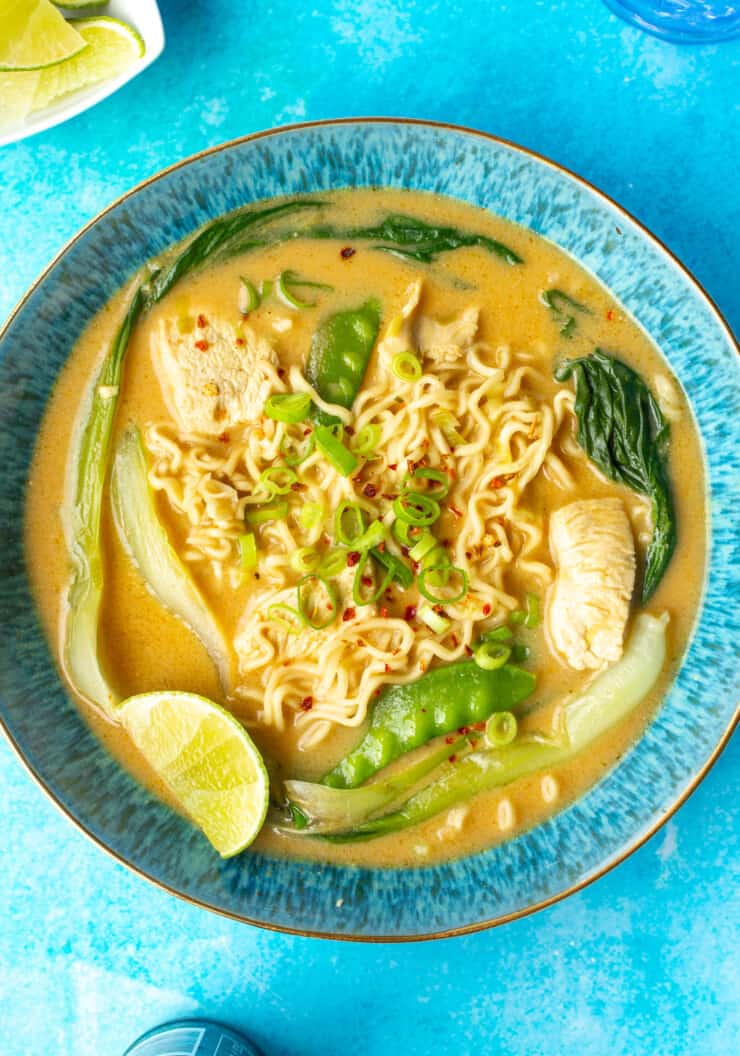 Chicken satay ramen with noodles, pak choi, mangetout, chicken, spring onion and a wedge lime in a blue bowl on a blue background.
