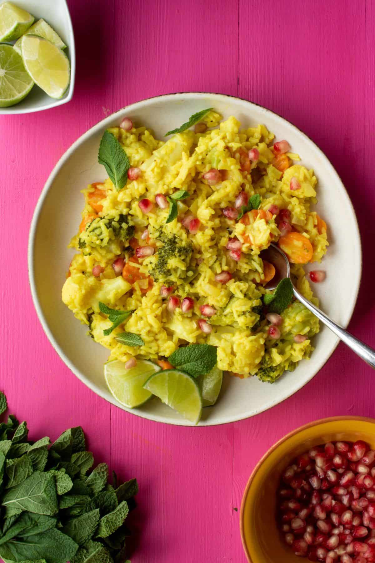 A bowl filled with yellow rice with fresh mint, vegetables, pomegranate and lime wedges with a spoon on a pink background next to some fresh mint, a bowl of pomegranate and lime wedges. 