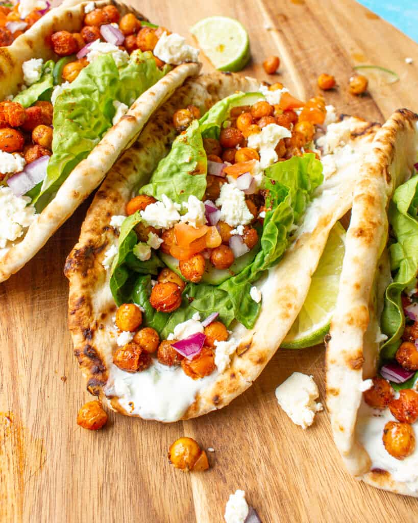 Chickpea gyros with tzatziki, roasted spiced chick peas, lettuce, feta on flat breads on a wooden board.