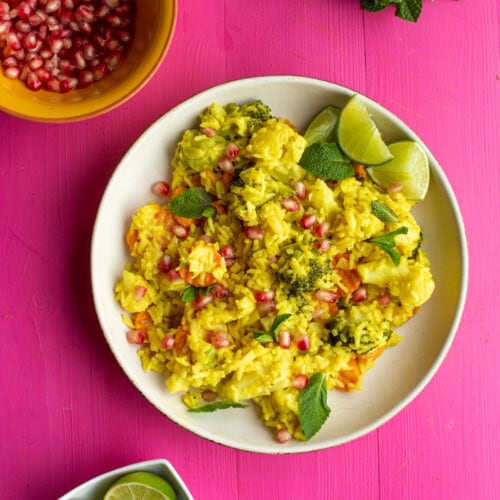 Golden Vegetable Pilaf rice in a white bowl on a pink background with small bowls of pomegranate and lime wedges and some chopped mint.