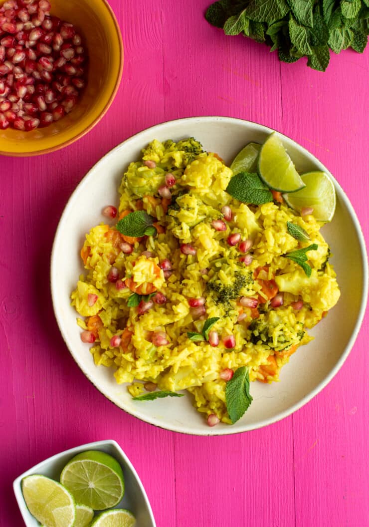 Golden Vegetable Pilaf rice in a white bowl on a pink background with small bowls of pomegranate and lime wedges and some chopped mint.