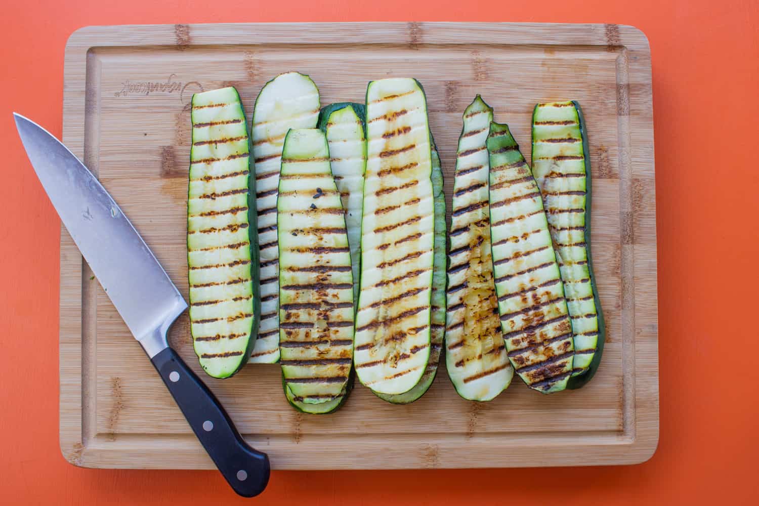 Roasted and charred courgette strips on a chopping board with a knife.