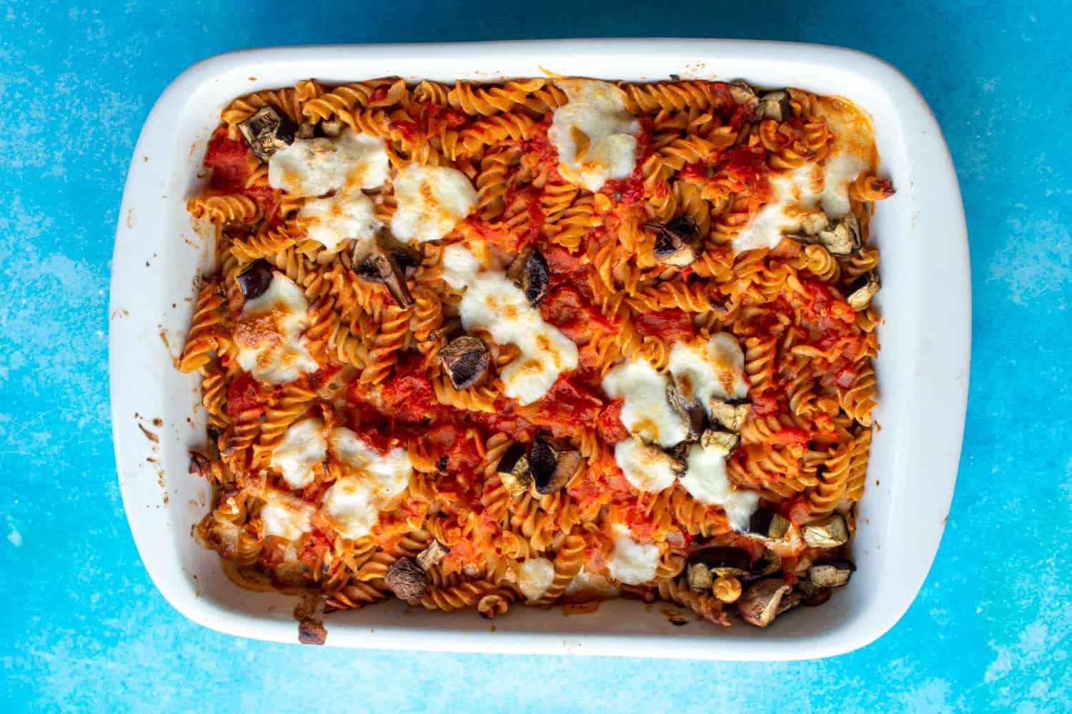Healthy Baked Ziti with fusilli pasta, aubergine, tomato sauce, mushrooms and browned mozzarella in white baking dish on a blue background.