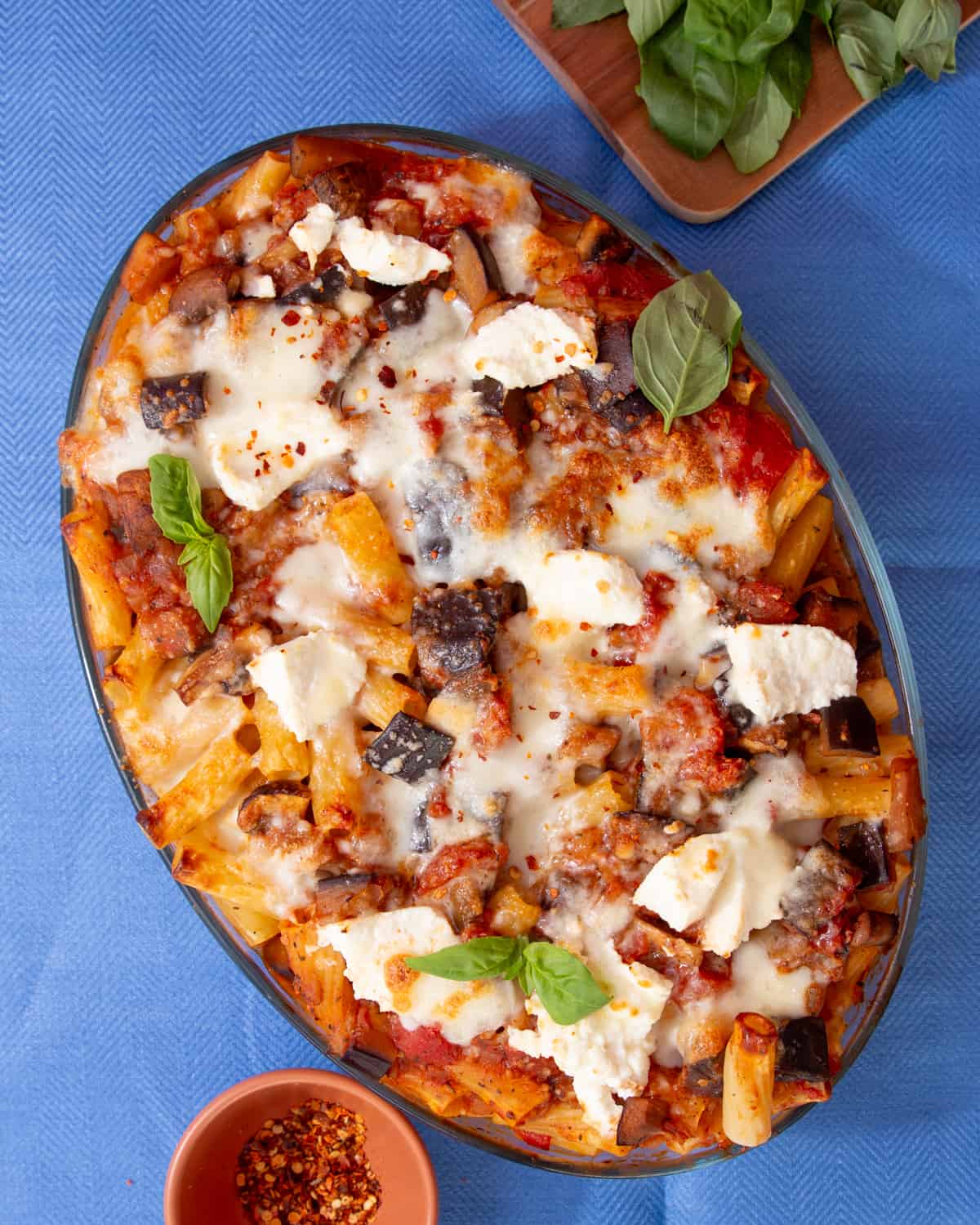 An oval dish filled with baked rigatoni pasta and a cheesy browned topping garnished with fresh basil on a blue background.