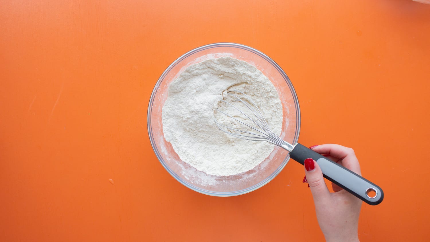 Flour and baking powder in a glass bowl and mixed with a metal whisk on an orange background.