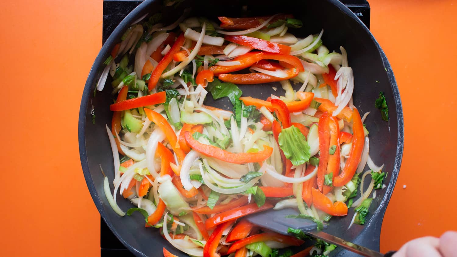 Sliced peppers, onions and pak choi in a frying pan mixed with a spatula on a cooker on an orange background.