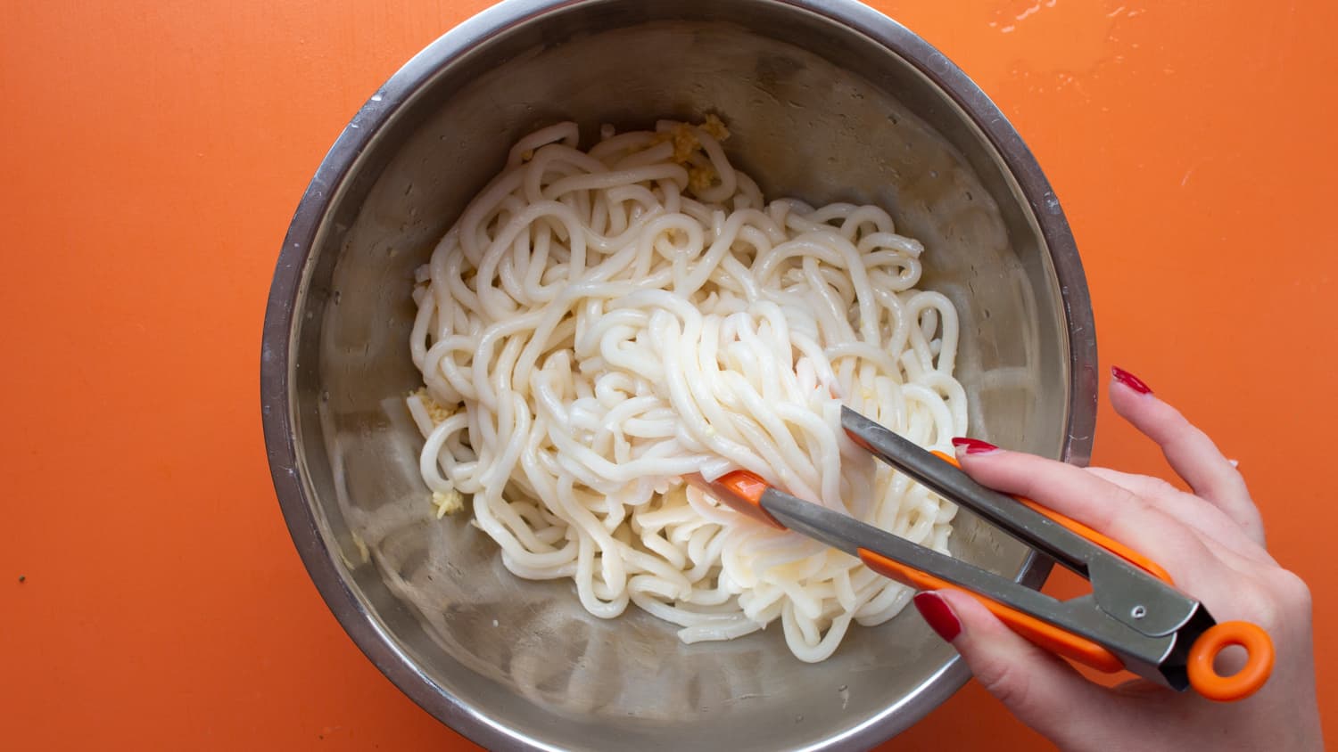Noodles ready in metal pan with minced garlic and chilli oil being mixed with orange tongs on an orange background.