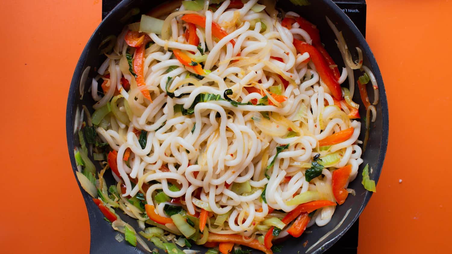 Udon noodles, sliced peppers, onions and pak choi mixed together in a frying pan on a cooker on an orange background.