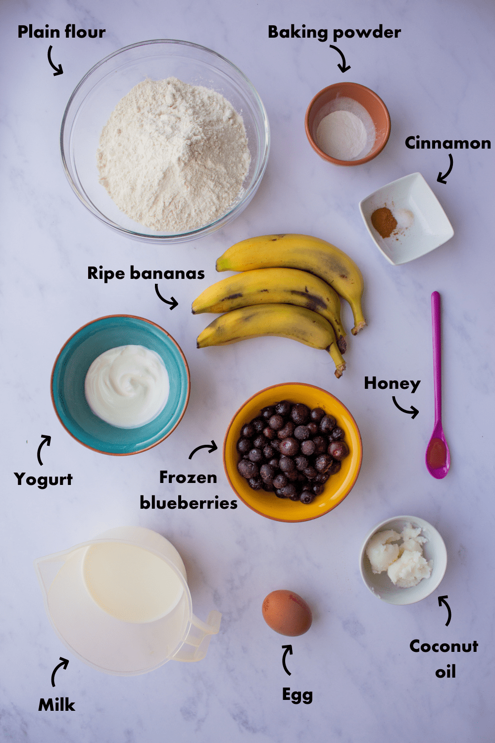 Ingredients to make Healthy Banana Blueberry Muffins laid out on a greyish background and labelled.