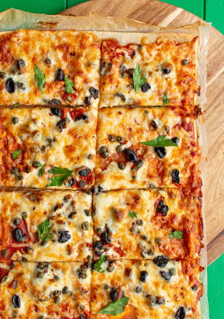 Sheet Pan Puttanesca Pizza with mozzarella, black olives, capers and fresh parsley cut into 8 pieces on parchment paper on a wooden board.
