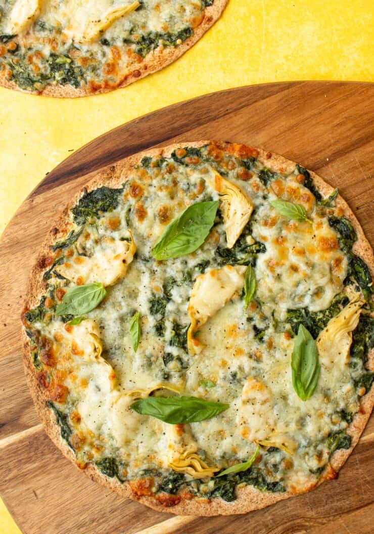 2 Spinach Artichoke Pizzas topped with mozzarella cheese, spinach and basil, one pizza on a wooden board, the other on a yellow background.