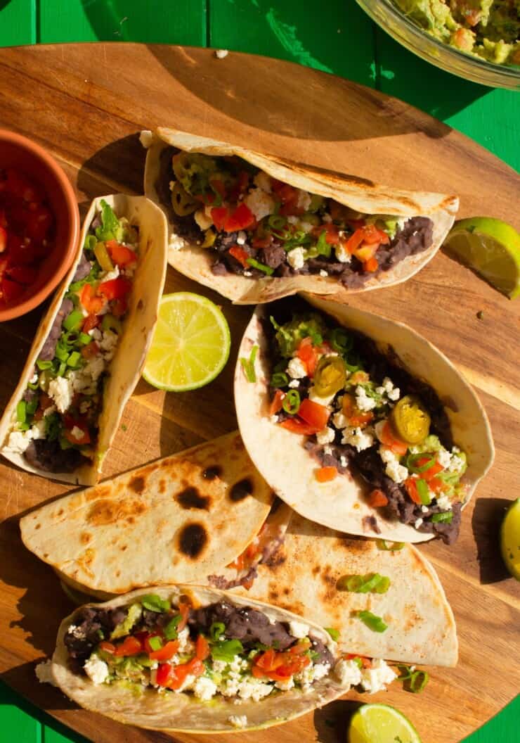 Black Bean Tacos with Guacamole with lime wedges, jalapeños and chopped tomatoes laid out on a wooden chopping board.