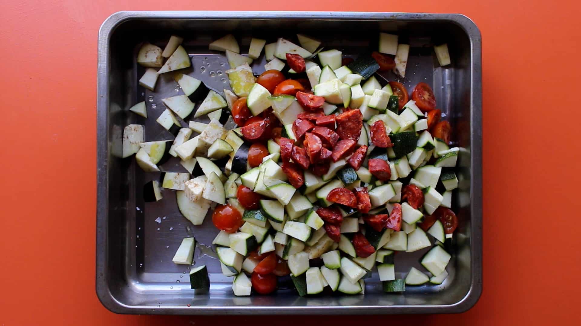 Chopped courgettes, tomatoes and sliced chorizo on a stainless steel baking tray on an orange background.