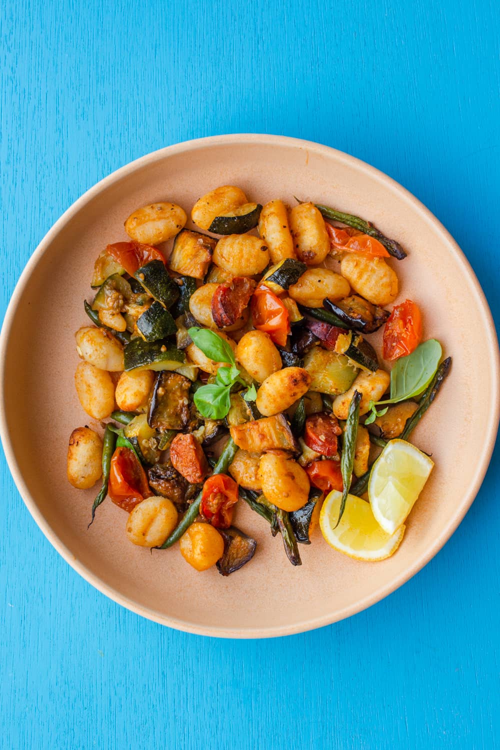 A bowl of Crispy Roasted Gnocchi with Chorizo and Vegetables with 2 wedges of lemon and fresh basil on a bright blue background.