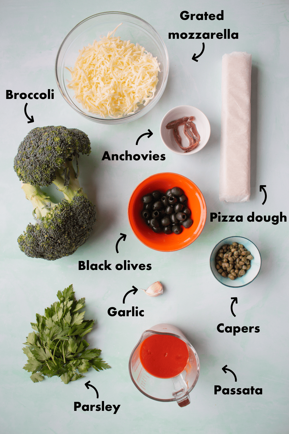 Ingredients to make Sheet Pan Puttanesca Pizza laid out on a grey background and labelled.