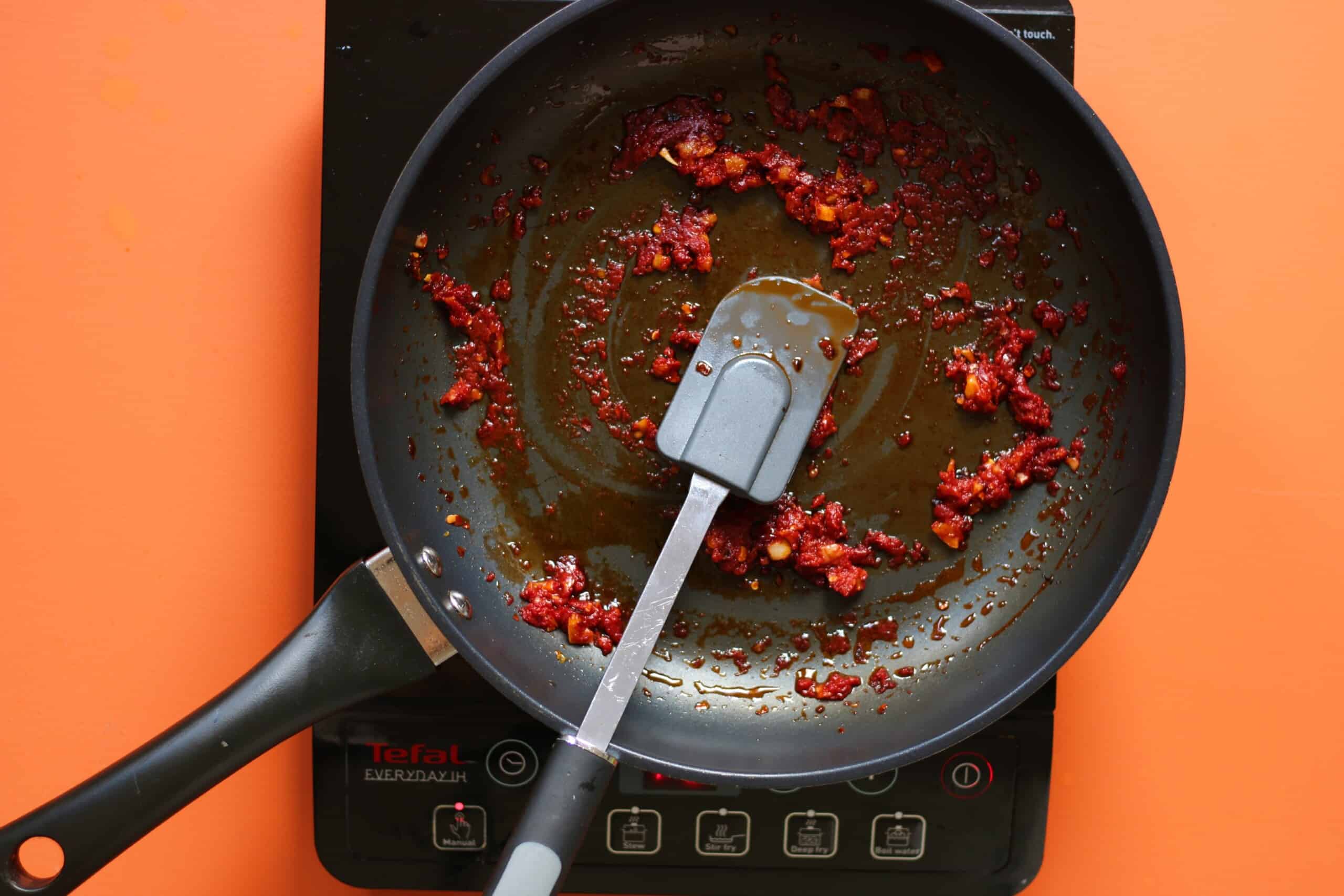 Fried tomato paste, chillies and garlic with oil in frying pan with spatula on a stove with an orange background.