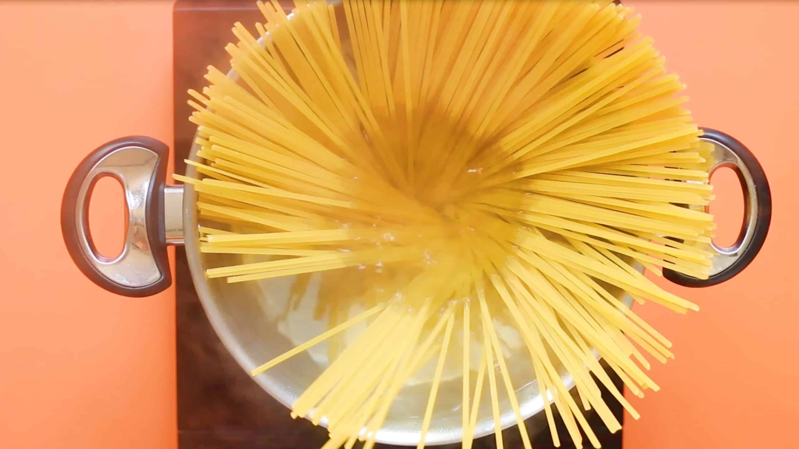 Dry spaghetti in sauce pan with 2 handles in water on stove on an orange background.
