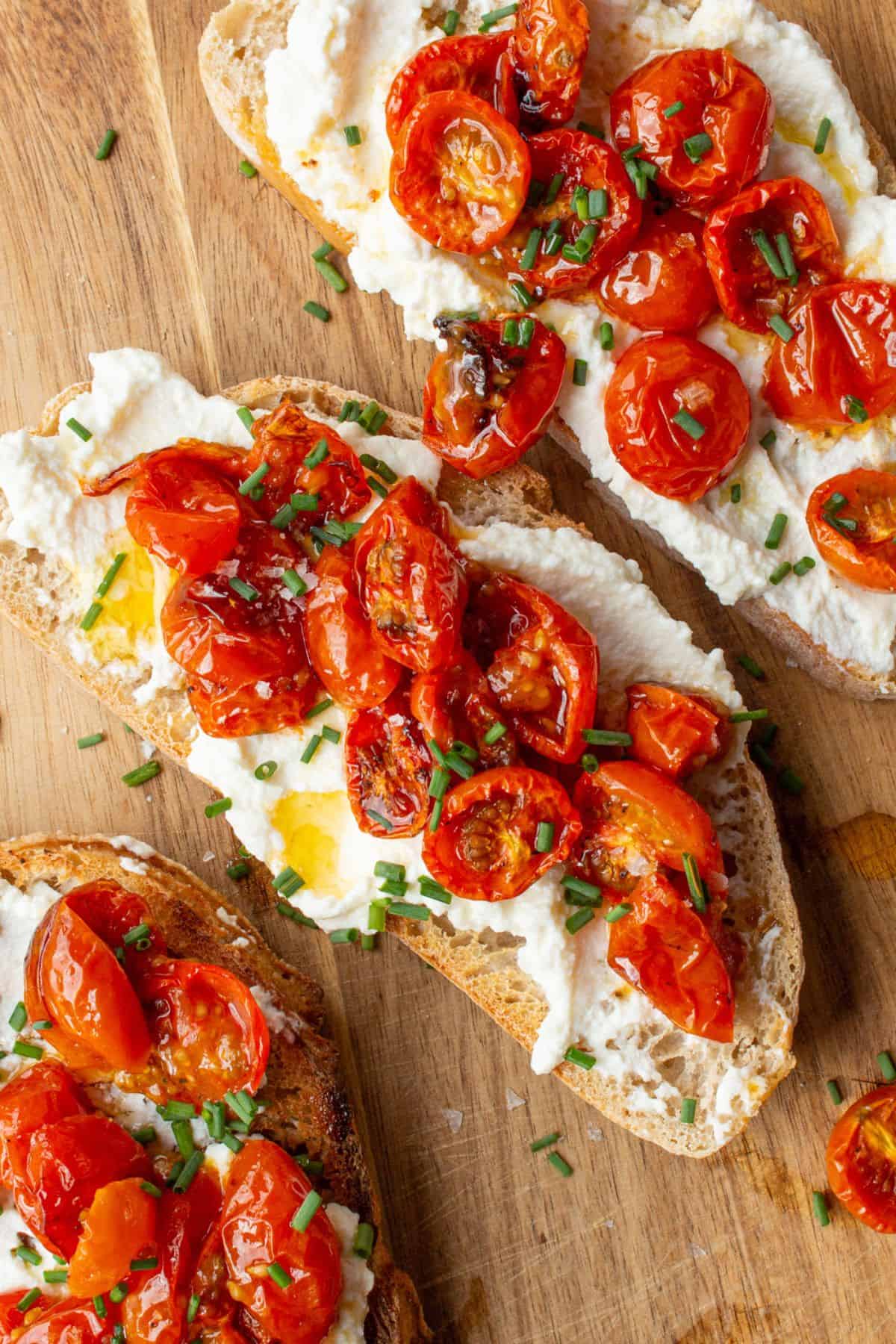 3 slices of sourdough bread with cream cheese and roasted cherry tomatoes topped with chives on a wooden background.